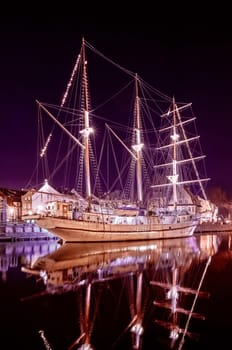 Sailing boat anchored in old town of Klaipeda, Lithuania, with evening lights on. Medieval town and barquentine reflection in calm waters.