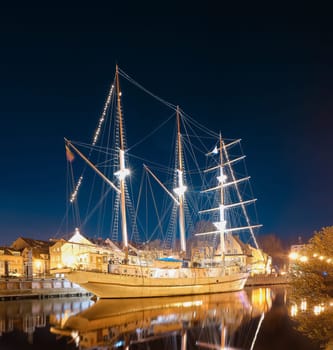 Sailing boat anchored in old town of Klaipeda, Lithuania, with evening lights on. Medieval town and barquentine reflection in calm waters.