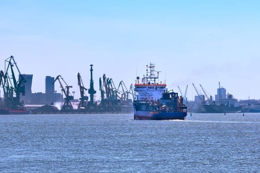 View of commercial cargo port and its container bridge cranes and a ship entering port on a sunny day.
