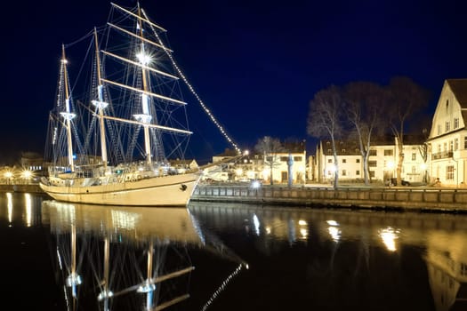 Sailing boat anchored in old town of Klaipeda, Lithuania, with evening lights on. Medieval town and barquentine reflection in calm waters.