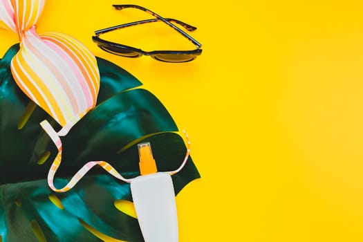 Tropical jungle palm monstera leaves, sunglasses, sunscreen blank bottle mockup and woman's swimsuit top on bright yellow paper background. Summer vacations creative flat lay concept template for text