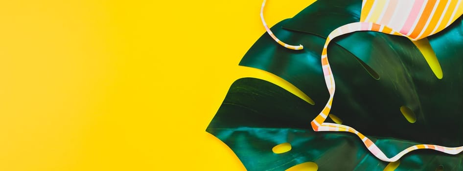 Tropical jungle palm monstera leaves and a woman's swimsuit top on bright yellow paper background. Summer vacations creative flat lay concept template for text. Long wide banner