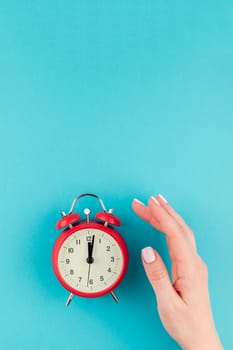 Creative flat lay concept top view of woman hand holding the red vintage alarm clock on bright blue turquoise color paper background with copy space in minimal style, template for text