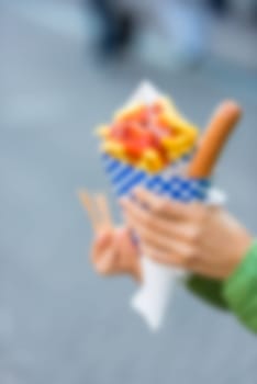 Female hands holding paper cone with a hot sausage and french fries with ketchup. Defocused shot