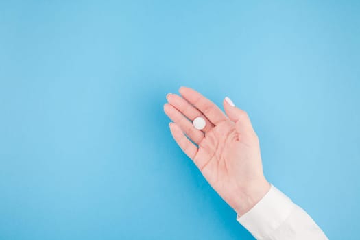 Woman hand holding a white pill on pastel blue paper background with copy space in minimal style, template for text. Concept of medical treatment, pharmacy, healthcare.