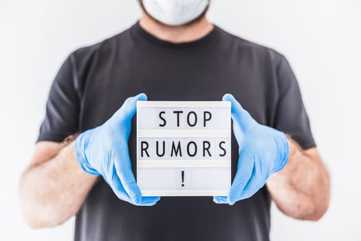 Fake news infodemics during Covid-19 pandemic concept. Man wearing protective mask and medical gloves on hands holding lightbox with text Stop rumors. People want to know the truth about coronavirus