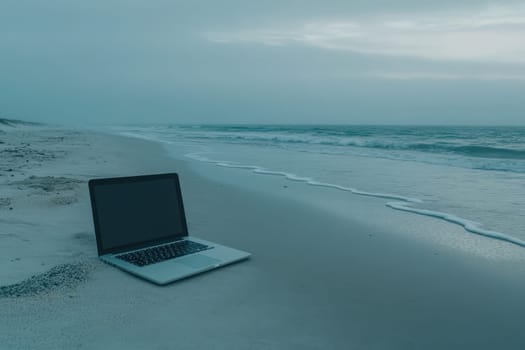 A sleek laptop is placed on a tranquil beach, with soft sand beneath and calm ocean waves lapping nearby. The scene captures a moment of peaceful solitude during twilight.