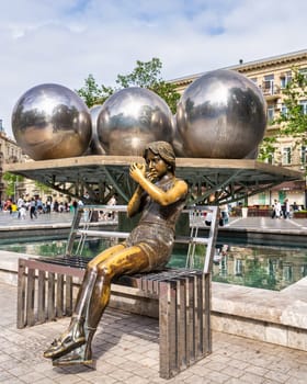Baku, Azerbaijan - May 5, 2024: Serene bronze statue depicts a girl sitting on a bench, lost in thought, situated in the Fountains Square in Nizami Street. The backdrop features striking metal spheres