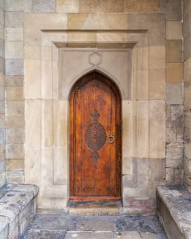 A beautifully crafted wooden door stands at the entrance of a historic stone building, showcasing intricate carvings and a unique arch. The surrounding walls are made of large, aged stones.