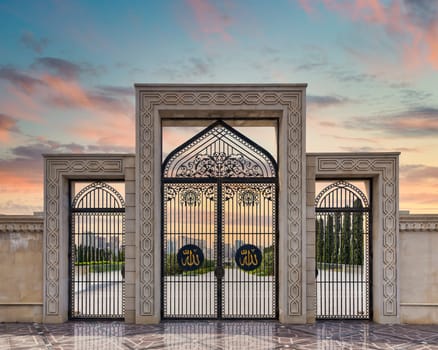 Ornate side gates of Taza Pir Mosque in Baku, Azerbaijan, glow under a colorful sunset, featuring two oval shaped metal signs with the Arabic word Allah, translates to God