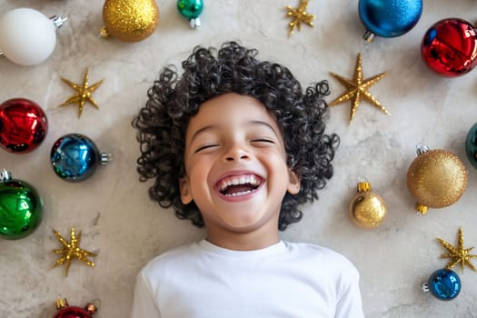 A cheerful young boy with curly hair laughs while lying on the floor amidst vibrant holiday decorations and ornaments.