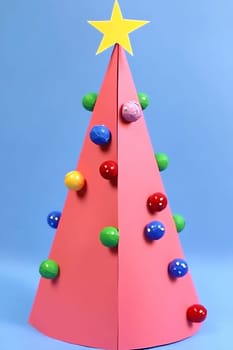 A handmade paper Christmas tree stands upright, adorned with colorful ornaments and topped with a bright star, set against a blue backdrop.