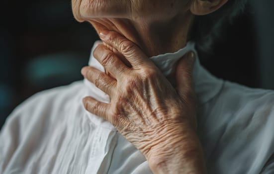 An older individual thoughtfully touches their throat, dressed in a white collared shirt, in a softly lit room.