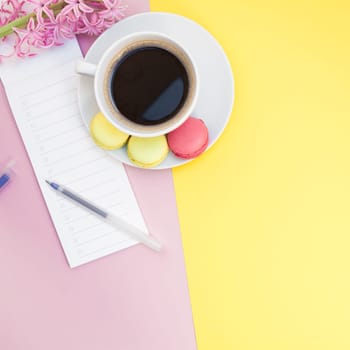 Creative square flat lay photo of coffee cup with macaroons and a notepad with copy space on pink and yellow background minimal style