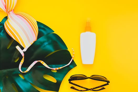Tropical jungle palm monstera leaves, sunglasses, sunscreen blank bottle mockup and woman's swimsuit top on bright yellow paper background. Summer vacations creative flat lay concept template for text