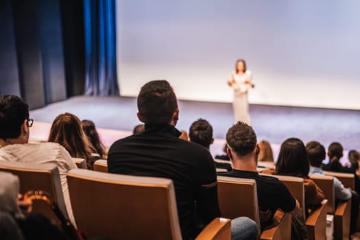 Business and entrepreneurship symposium. Female speaker giving a talk at business meeting. Audience in conference hall. Rear view of unrecognized participant in audience.