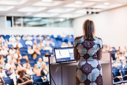 Female speaker giving a talk on corporate business conference. Unrecognizable people in audience at conference hall. Business and Entrepreneurship event.