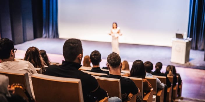 Business and entrepreneurship symposium. Female speaker giving a talk at business meeting. Audience in conference hall. Rear view of unrecognized participant in audience.