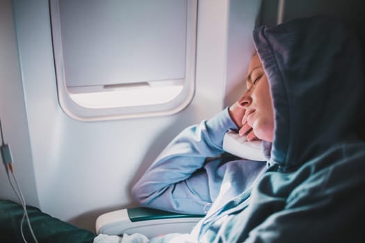 Tired blonde casual caucasian woman wearing sporty hoodie napping on seat while traveling by airplane. Commercial transportation by planes. Authentic image of real people