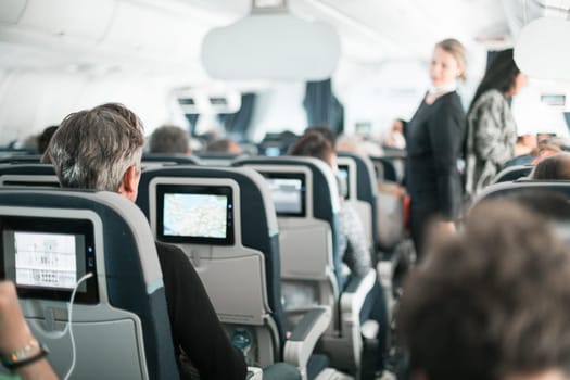 Interior of airplane with passengers on seats and stewardess in uniform walking the aisle, serving people. Commercial economy flight service concept