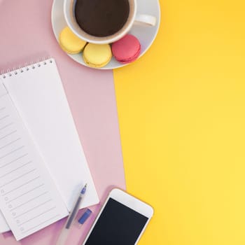 Creative square flat lay photo of coffee cup with macaroons and a notepad with copy space on pink and yellow background minimal style