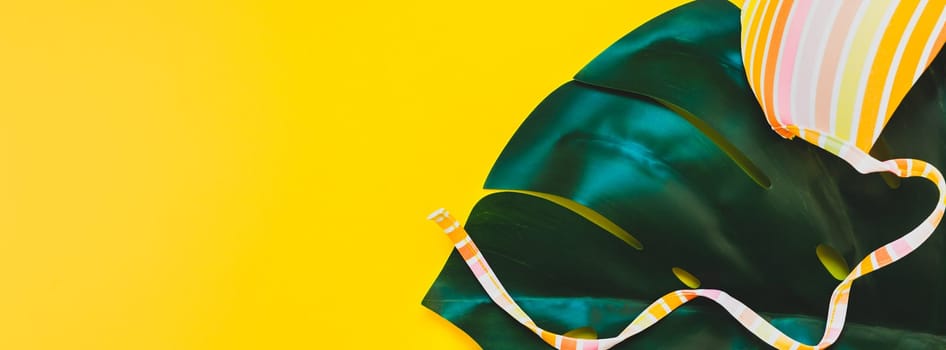 Tropical jungle palm monstera leaves and a woman's swimsuit top on bright yellow paper background. Summer vacations creative flat lay concept template for text. Long wide banner