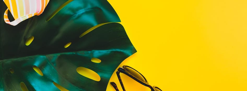 Tropical jungle palm monstera leaves, sunglasses and a woman's swimsuit top on bright yellow paper background. Summer vacations creative flat lay concept template for text. Long wide banner
