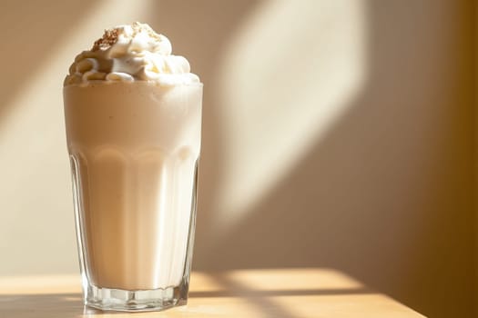 A tall, clear glass filled with a creamy milkshake is topped with whipped cream and a sprinkle of crumbs. Soft shadows play across a smooth wooden surface, enhancing the tranquil atmosphere.