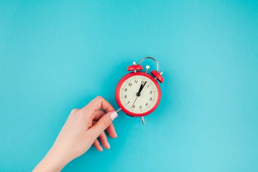 Creative flat lay concept top view of woman hand holding the red vintage alarm clock on bright blue turquoise color paper background with copy space in minimal style, template for text