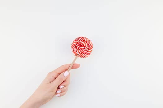 Woman hand with pastel manicure polish holding bright red swirling lollipop isolated on white background copy space minimalism style. Template for feminine social media. Fun and celebration concept