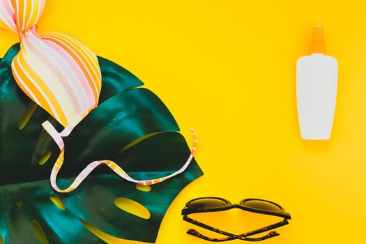 Tropical jungle palm monstera leaves, sunglasses, sunscreen blank bottle mockup and woman's swimsuit top on bright yellow paper background. Summer vacations creative flat lay concept template for text