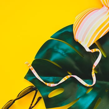 Tropical jungle palm monstera leaves, sunglasses and a woman's swimsuit top on bright square yellow paper background. Summer vacations creative flat lay concept template for text