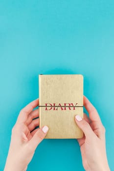 Creative flat lay of woman hands holding small golden notebook diary with copy space on turquoise paper background in minimalism style. Template for feminine blog, social media