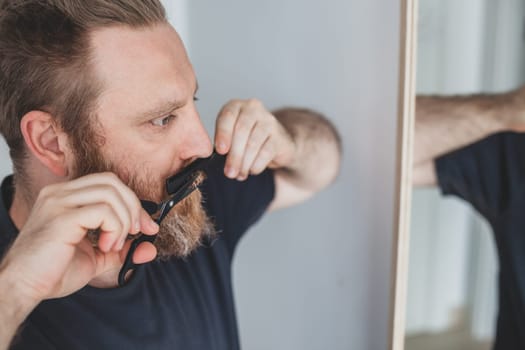 Selfcare during quarantine isolation. Adult handsome caucasian man cutting moustache and beard himself personally with scissors at home. Coronavirus outbreak and people shutdown, stay home concept