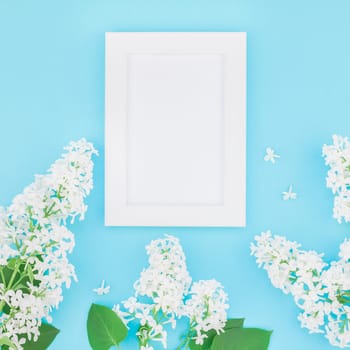 Square flat lay concept top view of blank postcard frame mock up and white lilac flowers petals on pastel blue background with copy space in minimal style, template for lettering, text or design