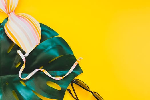 Tropical jungle palm monstera leaves, sunglasses and a woman's swimsuit top on bright yellow paper background. Summer vacations creative flat lay concept template for text