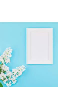 Creative flat lay concept top view of blank postcard frame mock up and white lilac flowers petals on pastel blue background with copy space in minimal style, template for lettering, text or design