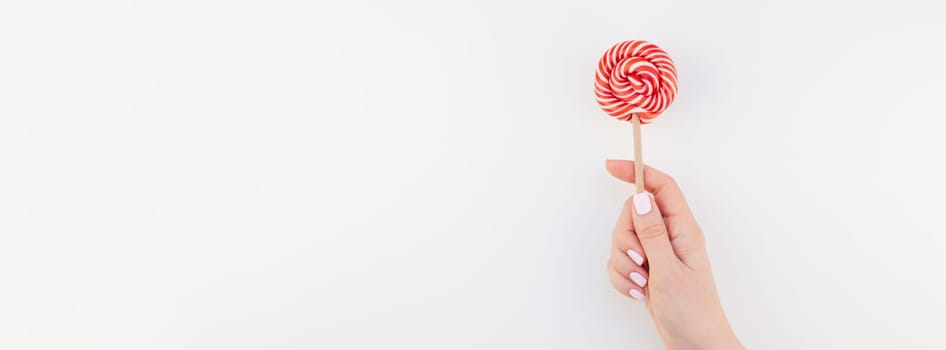 Woman hand pastel manicure polish holding bright red swirling lollipop isolated on white background copy space minimalism style. Long wide banner for feminine social media. Fun and celebration concept