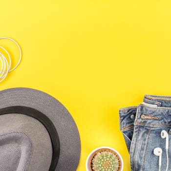 Flat lay of blue jeans, grey hat, cactus and smartphone headphones on bold yellow paper background with copy space. Overhead view of woman casual outfit. Square hipster look top view