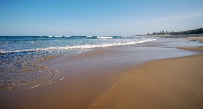 Popular tourist area, sandy beach with surf waves rolling in at Buddina, Sunshine Coast, Queensland, with clear, blue sky