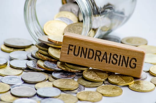 Fundraising text on wooden block with coins and jar background. Charity concept.
