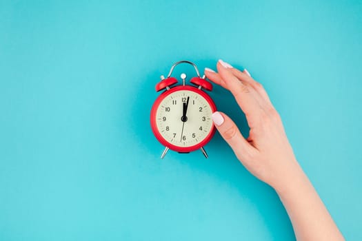 Creative flat lay concept top view of woman hand holding the red vintage alarm clock on bright blue turquoise color paper background with copy space in minimal style, template for text
