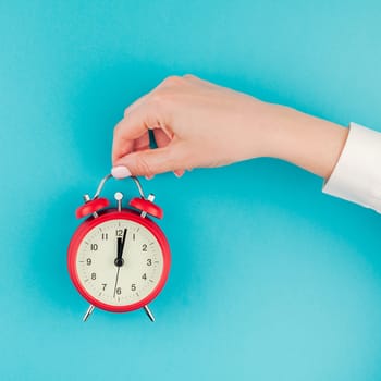 Creative flat lay concept top view of woman hand in white shirt holding the red vintage alarm clock on bright blue turquoise square color paper background with copy space in minimal style, template
