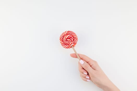 Woman hand with pastel manicure polish holding bright red swirling lollipop isolated on white background copy space minimalism style. Template for feminine social media. Fun and celebration concept