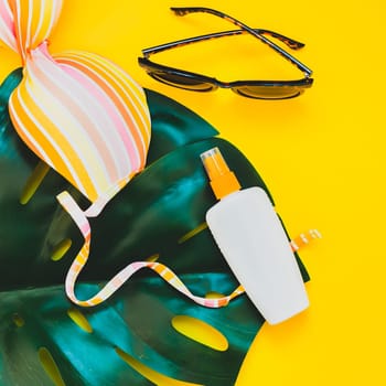 Tropical jungle palm monstera leaves, sunglasses, sunscreen blank bottle mockup and woman's swimsuit top on bright square yellow paper background. Summer vacations creative flat lay concept template