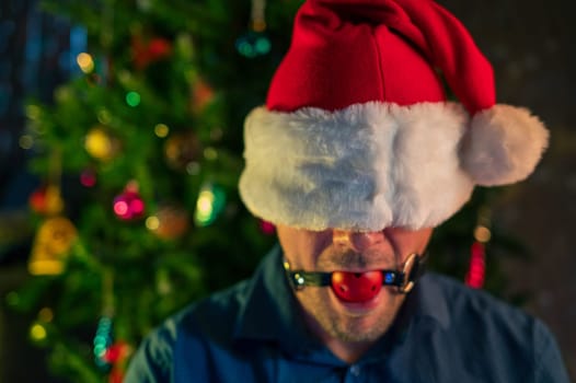 A man wearing a Santa hat, gagged, with a Christmas tree and festive lights in the background, provocative holiday scene