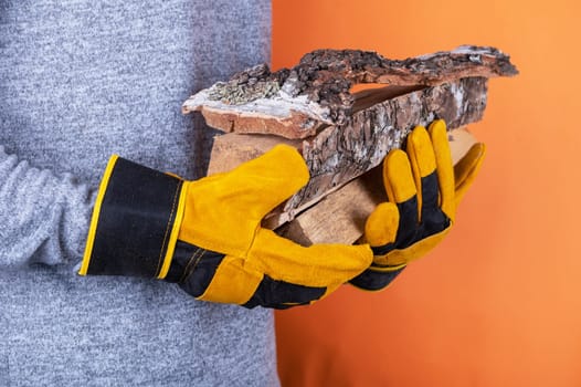 A person wearing yellow work gloves holds pieces of firewood against an orange background, emphasizing safety and outdoor work.