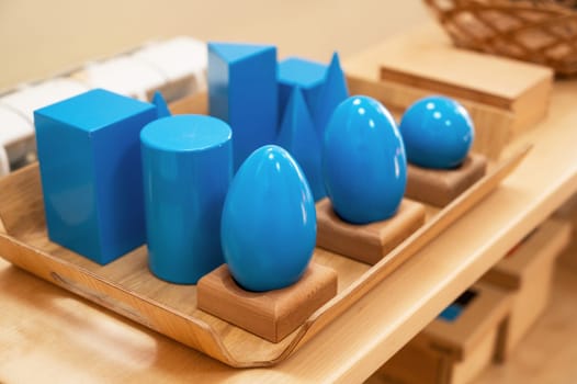 A set of blue geometric shapes, including spheres, cylinders, and cubes, neatly arranged on a wooden tray in a classroom.