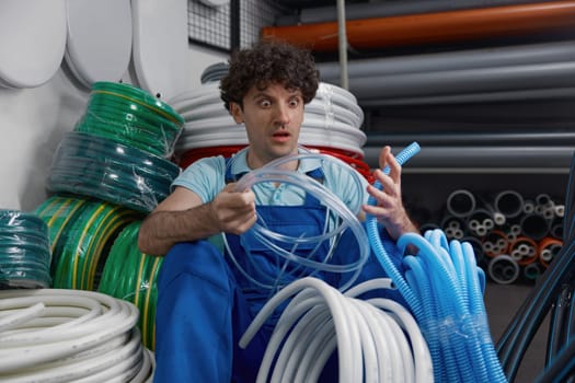 Crazy male plumber with mad facial expression confused in choice. Shouting while sitting at plumbing shop floor surrounded by variety and difference of water hoses and pipes