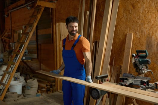 Young man carpenter in uniform putting wood plank on modern equipment closeup view. Experienced workman enjoying DIY woodworking project at his own small furniture factory